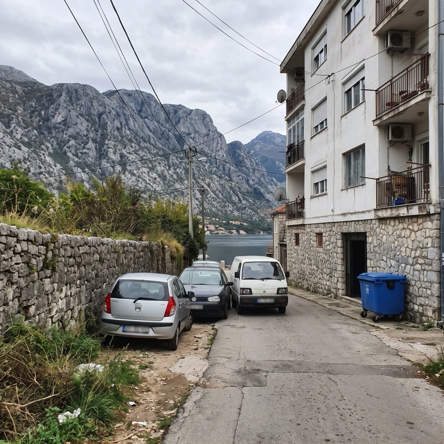 Narrow street in front of an apartment building in Dobrota with cars parked tightly along a stone wall and the bay in the background.